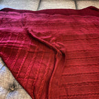 Claret Cotton-Blend Cable-Knit Throw: Red cable-knit throw with ribbed borders spread on a sofa.