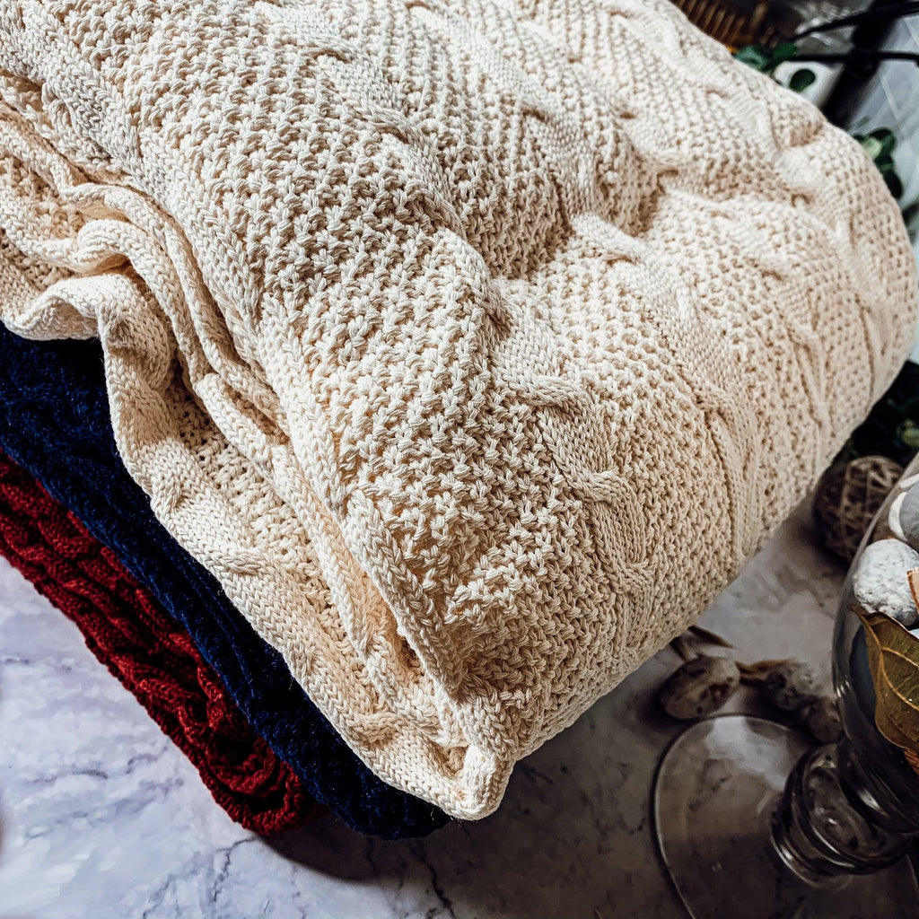 Claret Cotton-Blend Cable-Knit Throw: Folded cream cable-knit throw beside other colored throws.
