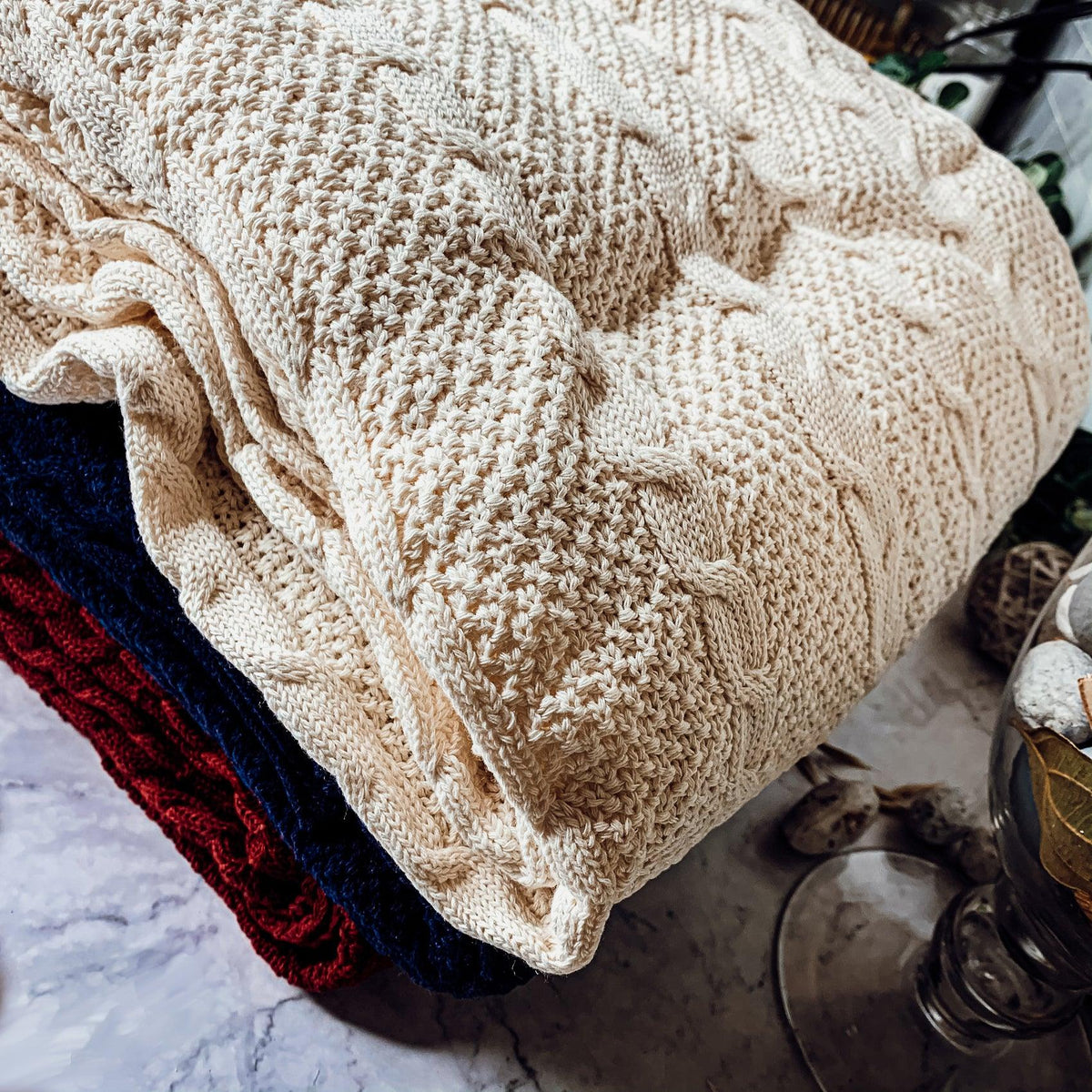 Claret Cotton-Blend Cable-Knit Throw: Folded cream cable-knit throw beside other colored throws.