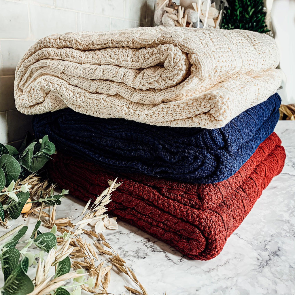 Claret Cotton-Blend Cable-Knit Throw: Stack of cream, blue and red cable-knit throws.