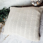 Claret Cotton-Blend Cable-Knit Throw: Cream cable-knit throw spread beside greenery.