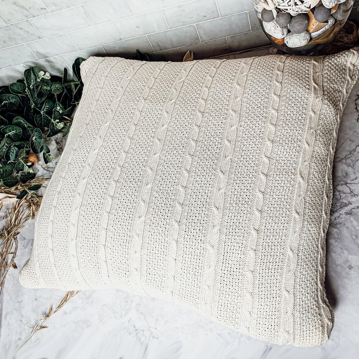 Claret Cotton-Blend Cable-Knit Throw: Cream cable-knit throw spread beside greenery.