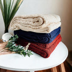 Claret Cotton-Blend Cable-Knit Throw: Stack of cream, blue and red throws on a round white table.